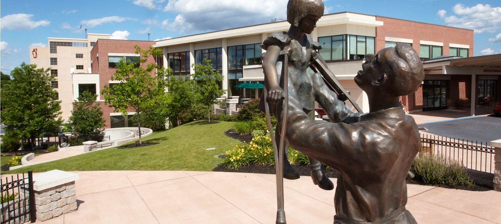 A photo of the Hyland Center for Health & Technology. In the foreground is a statue of "Papa Raker" holding a child with crutches in his arms. He holds her above his head.