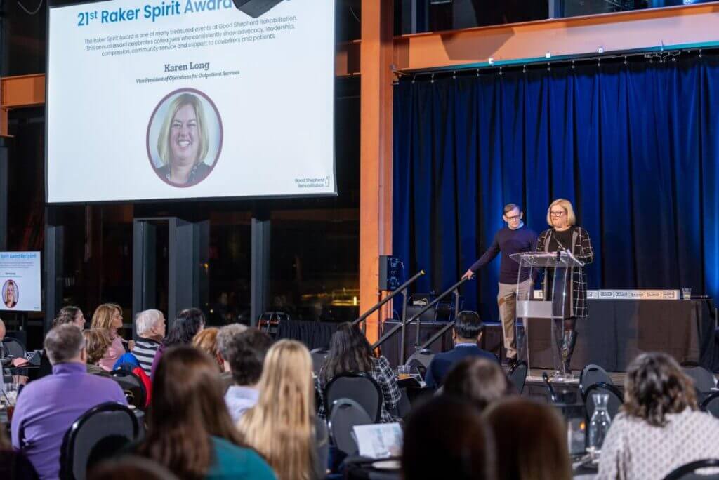 Karen Long stands at a podium and delivers a speech while Michael Spigel looks on to her right