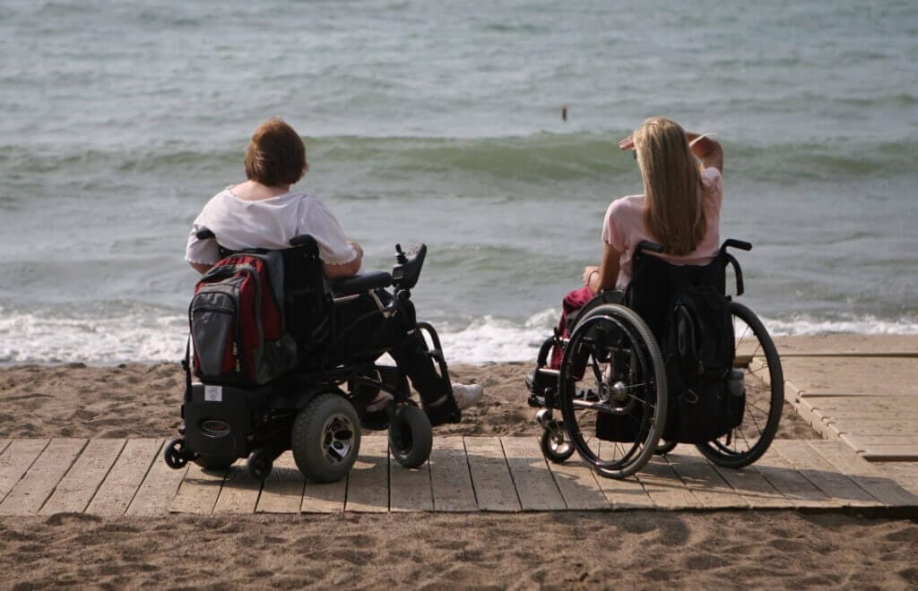 Two friends in wheelchairs on the boardwalk look out at the ocean
