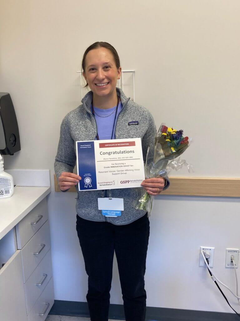 A person wearing a grey sweat-jacket holds a certificate that reads "Congratulations" and a bouquet of flowers.