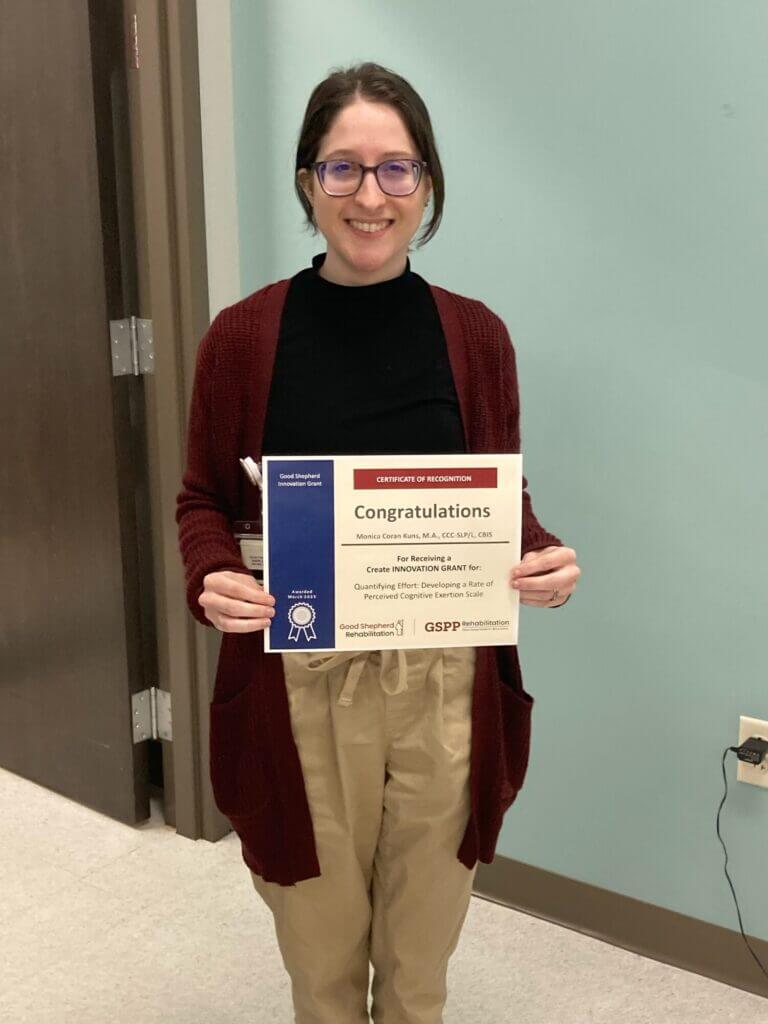 A person in a red cardigan and black turtleneck smiles while holding a certificate that reads "Congratulations"