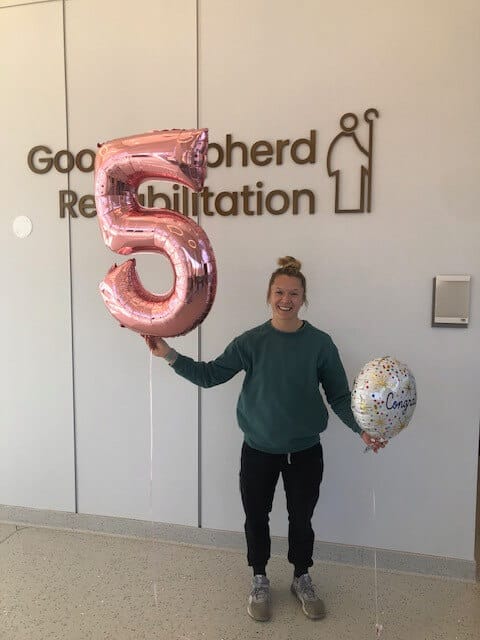 A person in a sweatshirt holds two balloons, one that says "Congratulations" and another of the number 5