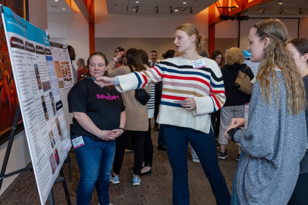 A group pf people talk to each other in a crowded room. One person is pointing to a poster presentation while talking to the others.
