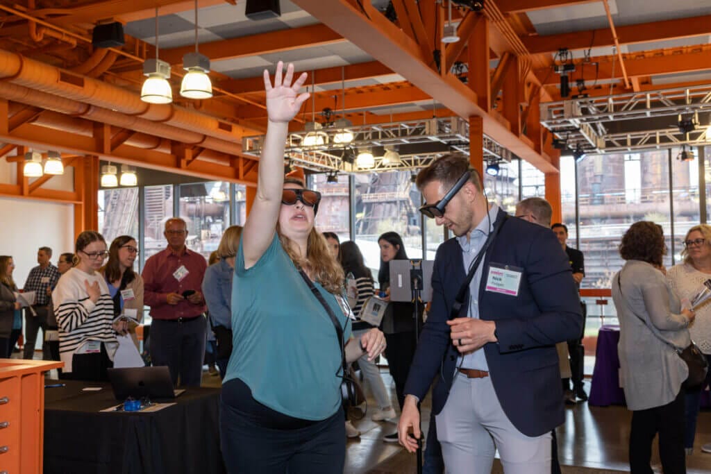 Two people are using AR Technology in a crowded room. The person on the left is wearing AR goggles while reaching upwards. The other is wearing the same set but looking downwards.