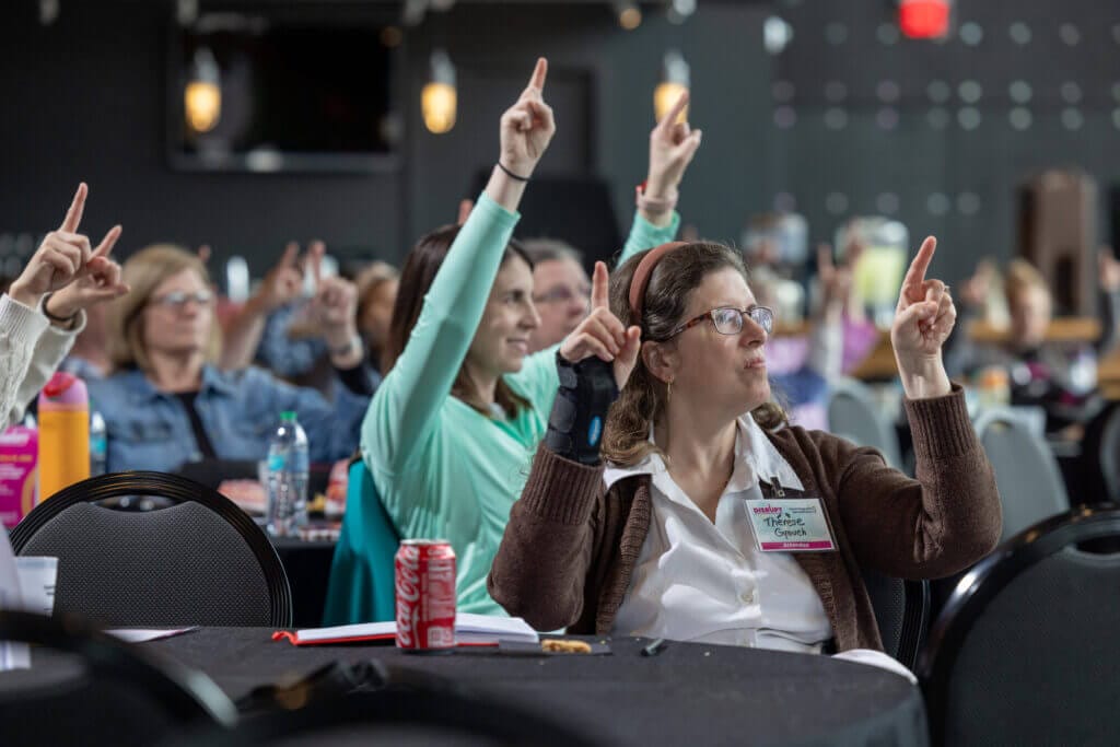 A group of people are holding both arms upwards, pointing with their index fingers.