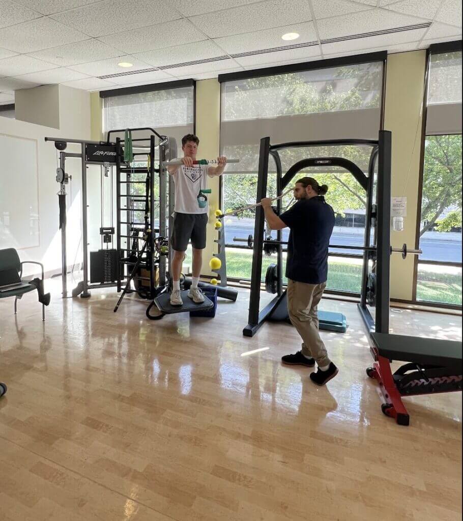 Noah and his Physical Therapist in a gym, one standing on a workout machine, the other assisting. Bright, naturally lit room with gym equipment and windows overlooking trees.
