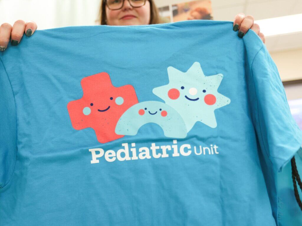 A Good Shepherd Rehabilitation pediatric hospital team member holds a light blue T-shirt that features colorful characters and the words "Pediatric Unit" on it.
