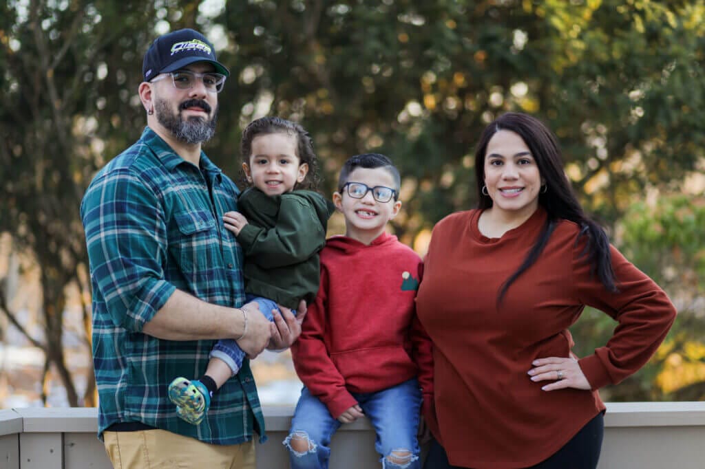 A family of four stands outside and poses for a family photo, smiling